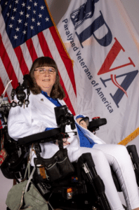 A woman in a white jacket and blue shirt sits in a motorized wheelchair. She is smiling and has long brown hair and glasses. Behind her are the American flag and a banner that reads 